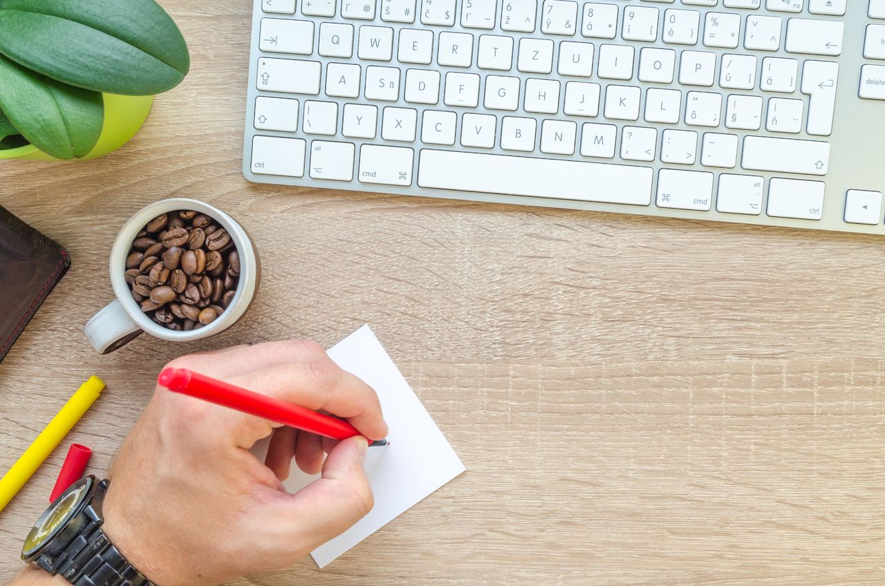 about-us A workspace featuring a keyboard, coffee beans, note, and a hand writing with a red pen.