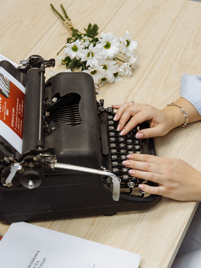 who-we-are A vintage typewriter with a woman's hands typing, surrounded by flowers on a wooden table.